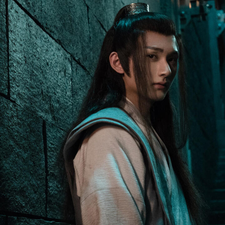 nie huaisang stands with his back against the wall, staring out into the camera.